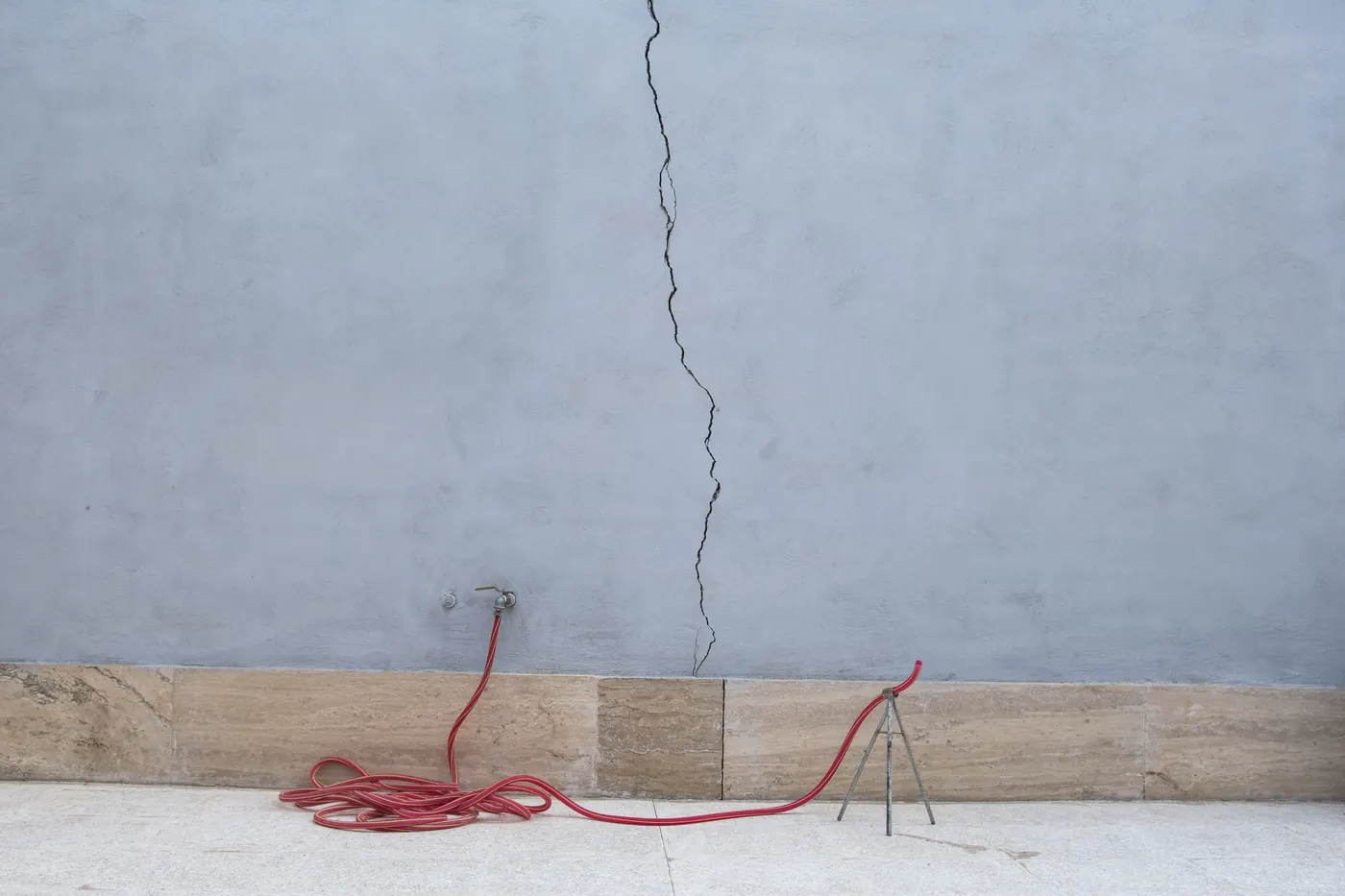 A long vertical crack running down a gray plastered wall to a stone base, with a red garden hose coiled below